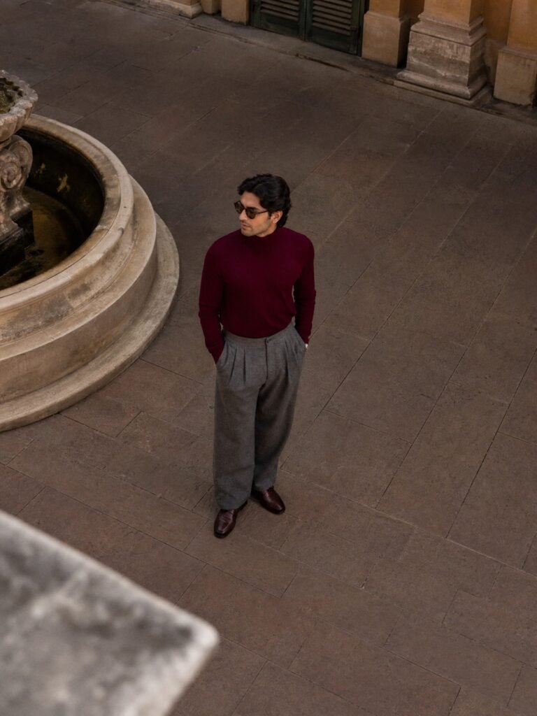 A man in wide-leg pleated grey wool trousers with a tucked-in merlot roll-neck and loafers