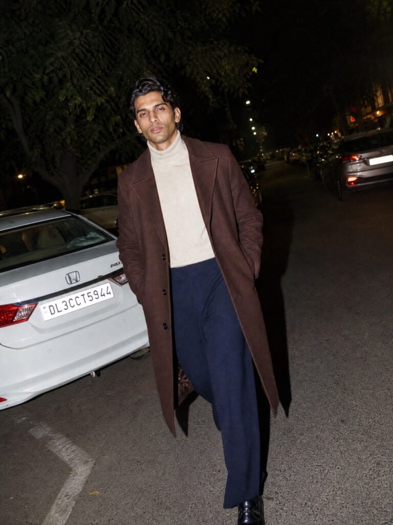 A man in a long brown overcoat, roll-neck and wide trousers on a winter street