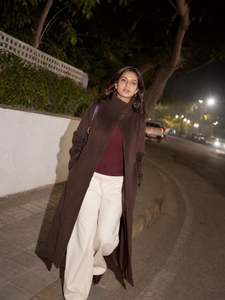 A woman in a long brown funnel-neck coat, burgundy knit and wide cream trousers on a winter street