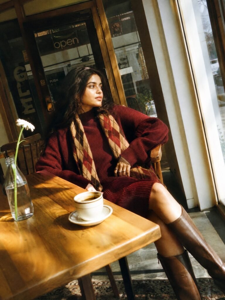 A woman in a long burgundy sweater dress with broad shoulders, an argyle scarf and knee-high brown boots
